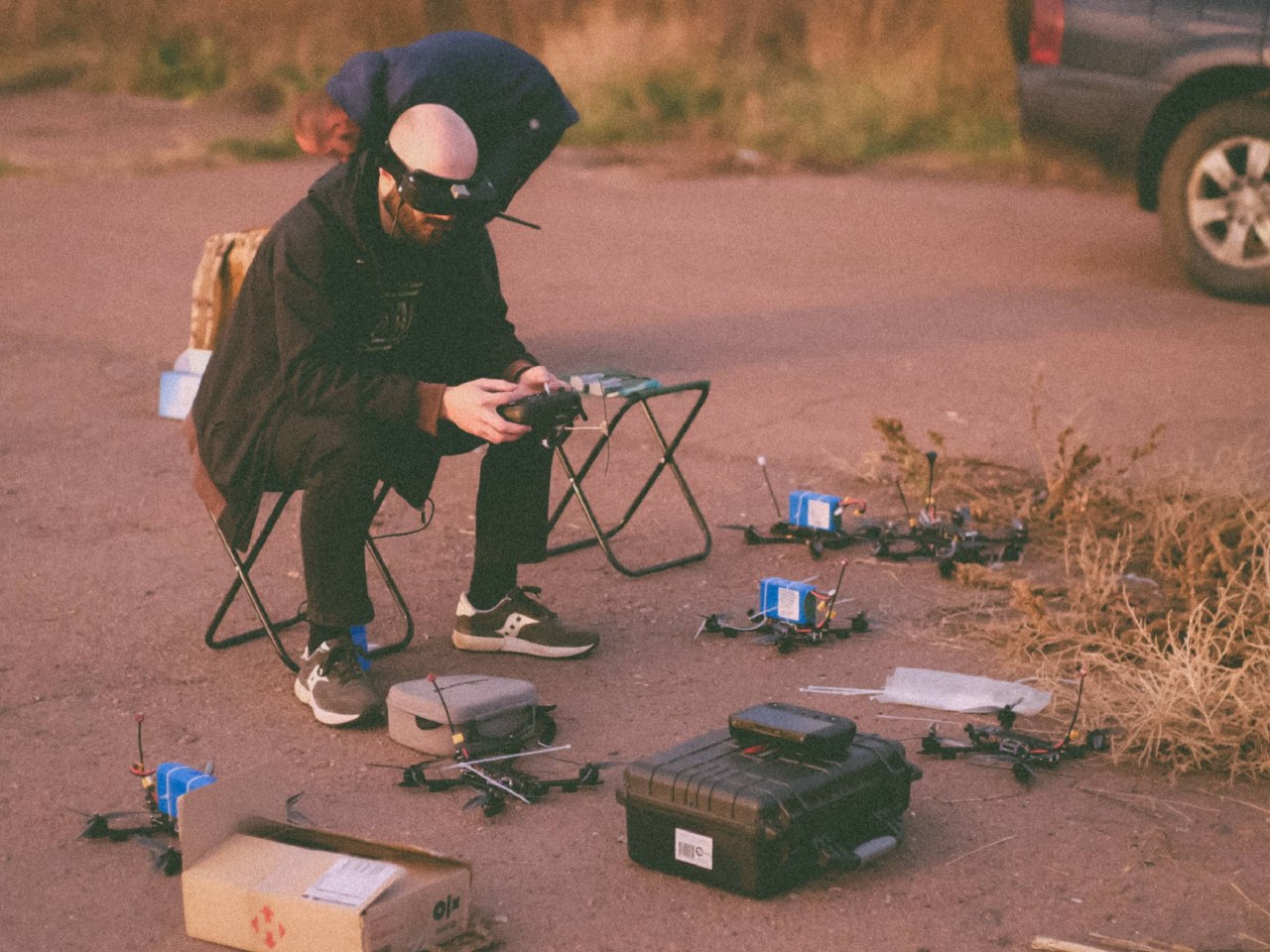 Operator seated outdoors in fading light, controller in hand, with several FPV airframes, batteries and equipment cases laid out on the ground